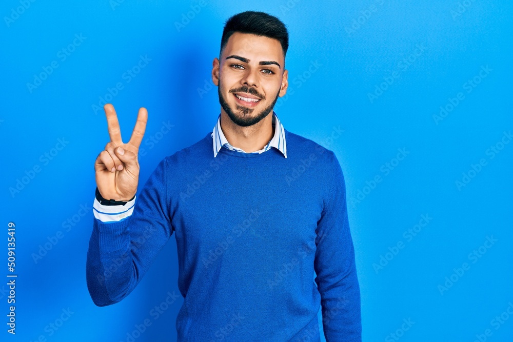 Fototapeta premium Young hispanic man with beard wearing casual blue sweater showing and pointing up with fingers number two while smiling confident and happy.