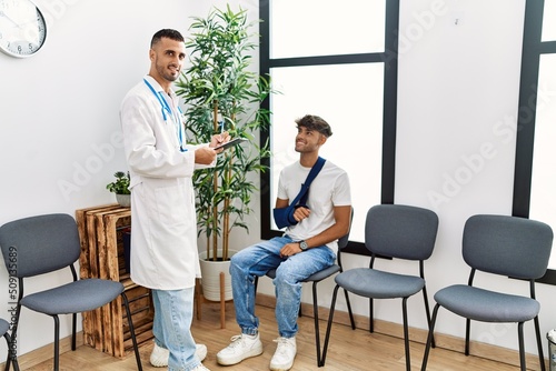 Two hispanic men doctor and patient writing on clipboard sitting on chair at hospital waiting room