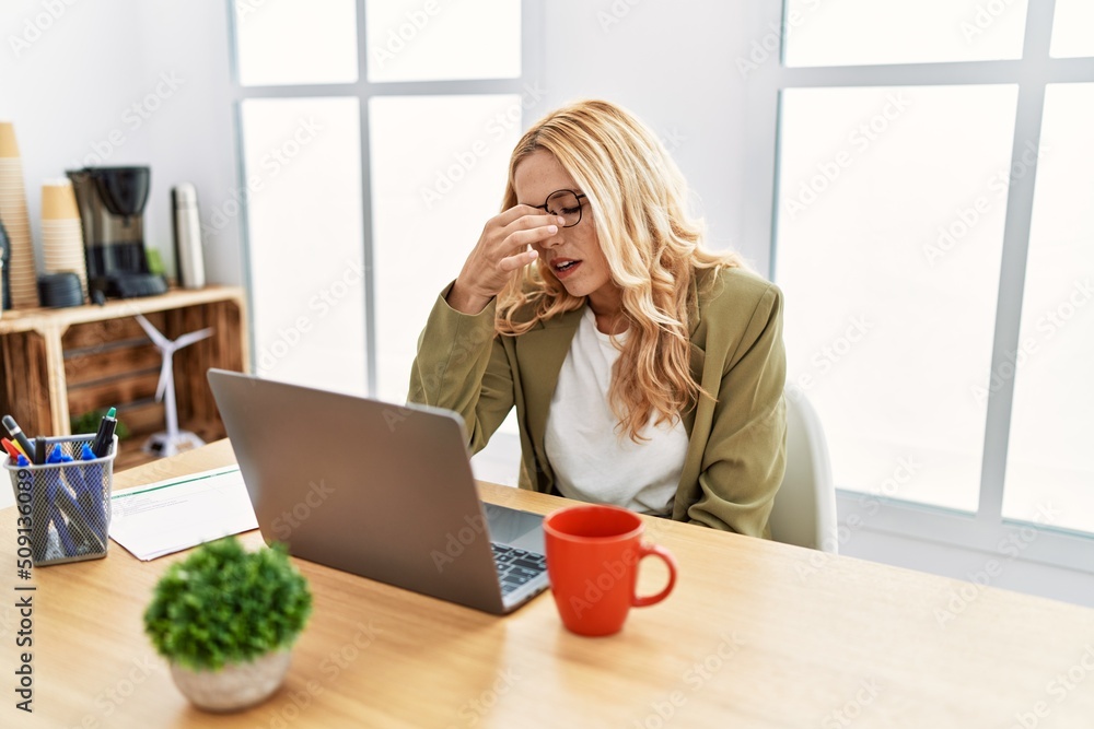 Beautiful blonde woman working at the office with laptop tired rubbing nose and eyes feeling fatigue and headache. stress and frustration concept. Beautiful blonde woman working at the office with laptop tired rubbing nose and eyes feeling fatigue and headache. stress and frustration concept.