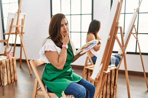 Young hispanic artist women painting on canvas at art studio looking confident with smile on face, pointing oneself with fingers proud and happy.