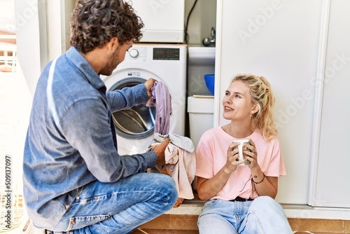Young couple smiling happy drinking coffee while doing laundry at home.