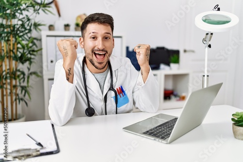 Young doctor working at the clinic using computer laptop celebrating surprised and amazed for success with arms raised and open eyes. winner concept.