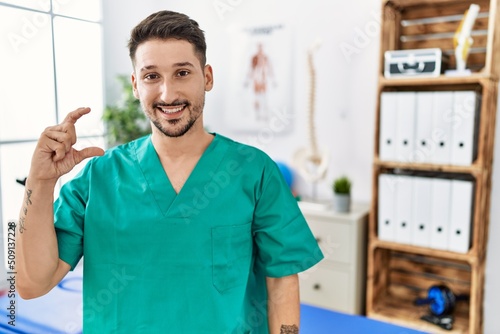 Young physiotherapist man working at pain recovery clinic smiling and confident gesturing with hand doing small size sign with fingers looking and the camera. measure concept.
