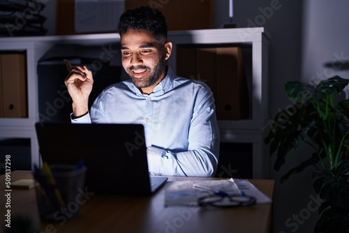 Hispanic man with beard working at the office with laptop at night smiling happy pointing with hand and finger to the side