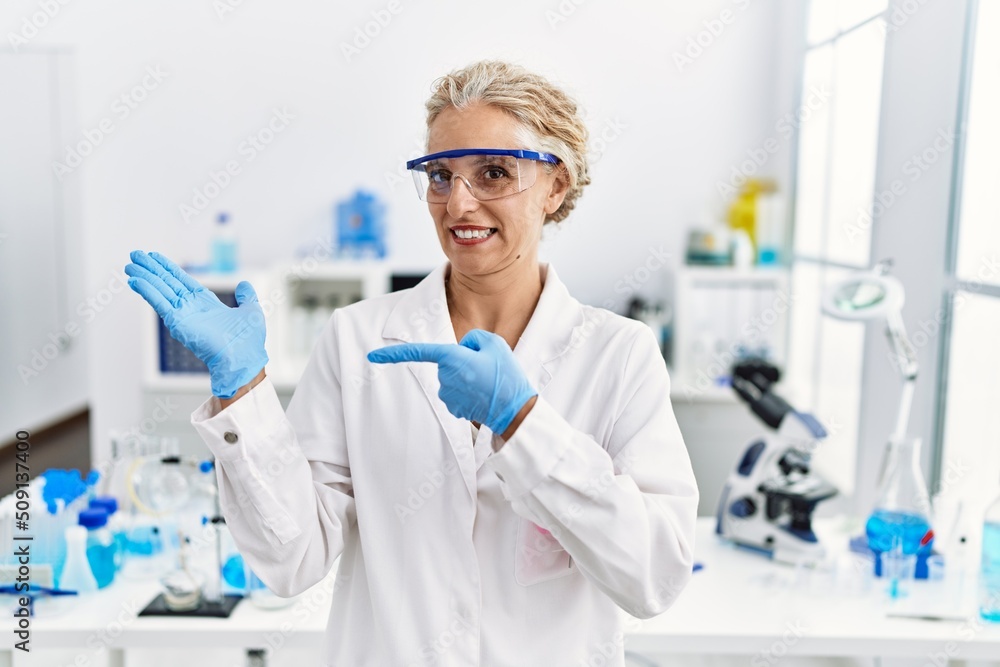 Middle age blonde woman working at scientist laboratory amazed and smiling to the camera while presenting with hand and pointing with finger. Middle age blonde woman working at scientist laboratory amazed and smiling to the camera while presenting with hand and pointing with finger.