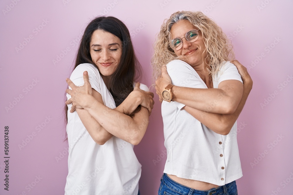 Mother and daughter standing together over pink background hugging oneself happy and positive, smiling confident. self love and self care Mother and daughter standing together over pink background hugging oneself happy and positive, smiling confident. self love and self care