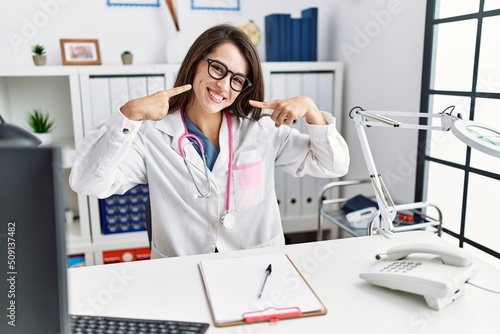 Young doctor woman wearing doctor uniform and stethoscope at the clinic smiling cheerful showing and pointing with fingers teeth and mouth. dental health concept.