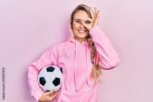 Beautiful young blonde woman holding soccer ball smiling happy doing ok sign with hand on eye looking through fingers