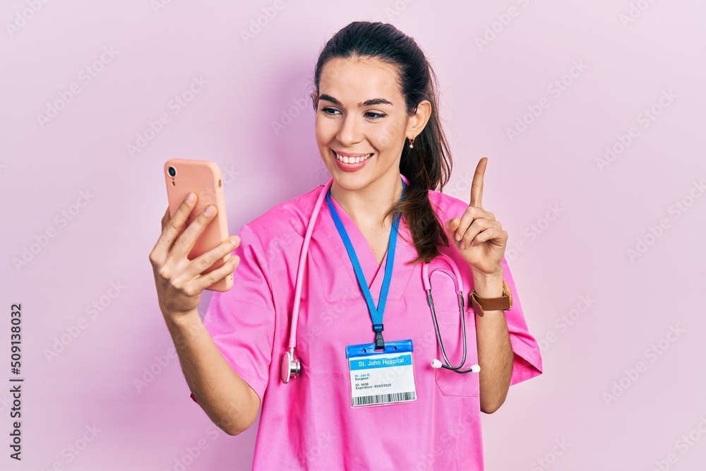 Young brunette doctor woman doing video call consult with smartphone smiling with an idea or question pointing finger with happy face, number one Young brunette doctor woman doing video call consult with smartphone smiling with an idea or question pointing finger with happy face, number one