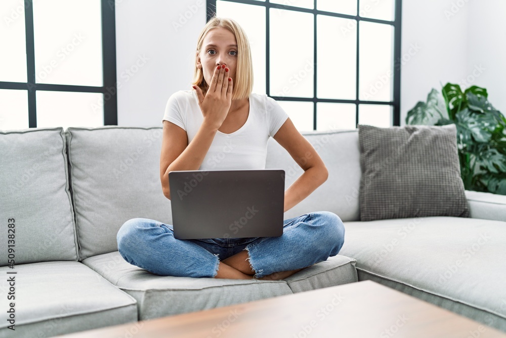 Young caucasian woman using laptop at home sitting on the sofa covering mouth with hand, shocked and afraid for mistake. surprised expression Young caucasian woman using laptop at home sitting on the sofa covering mouth with hand, shocked and afraid for mistake. surprised expression