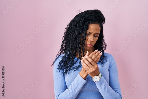 Middle age hispanic woman standing over pink background suffering pain on hands and fingers, arthritis inflammation