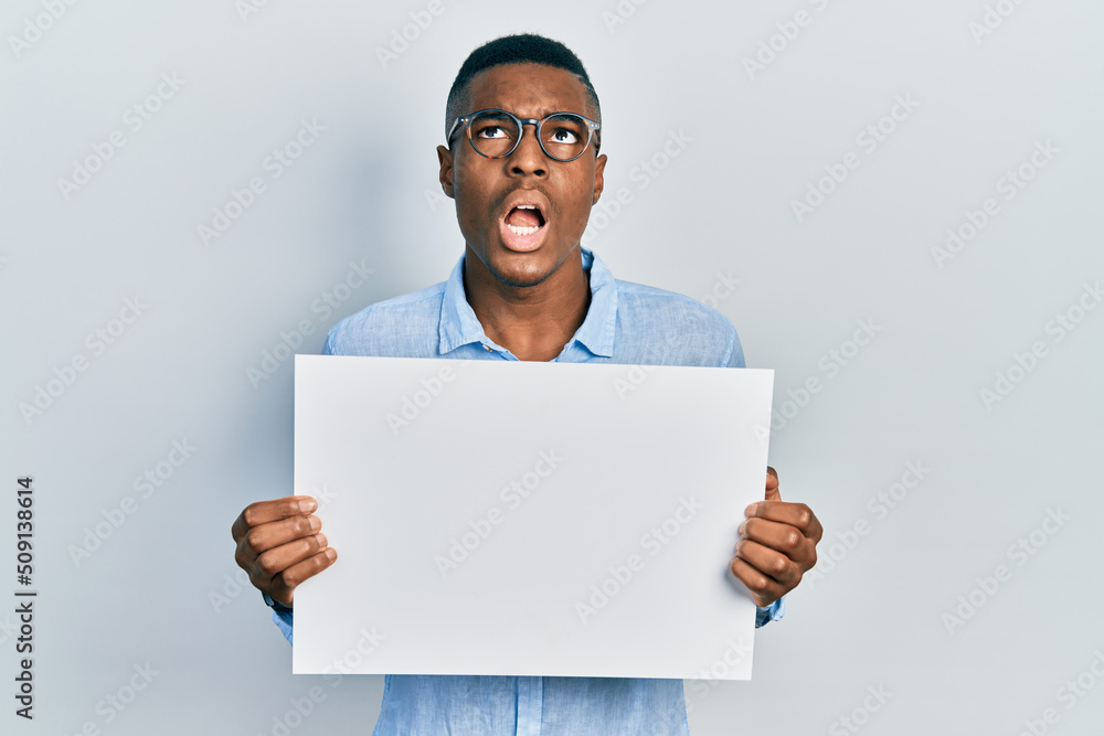 Young african american man wearing glasses holding blank empty banner angry and mad screaming frustrated and furious, shouting with anger looking up. Young african american man wearing glasses holding blank empty banner angry and mad screaming frustrated and furious, shouting with anger looking up.