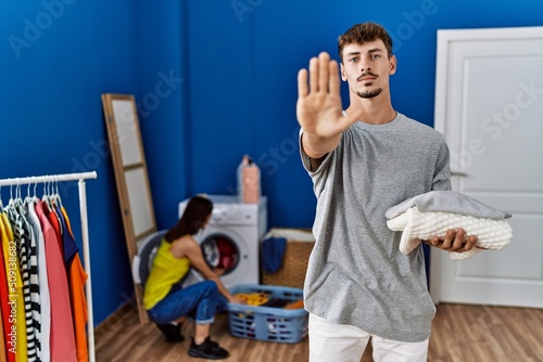 Young handsome man holding folded laundry with open hand doing stop sign with serious and confident expression, defense gesture © Krakenimages.com
