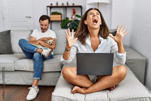 Hispanic middle age couple at home, woman using laptop crazy and mad shouting and yelling with aggressive expression and arms raised. frustration concept.
