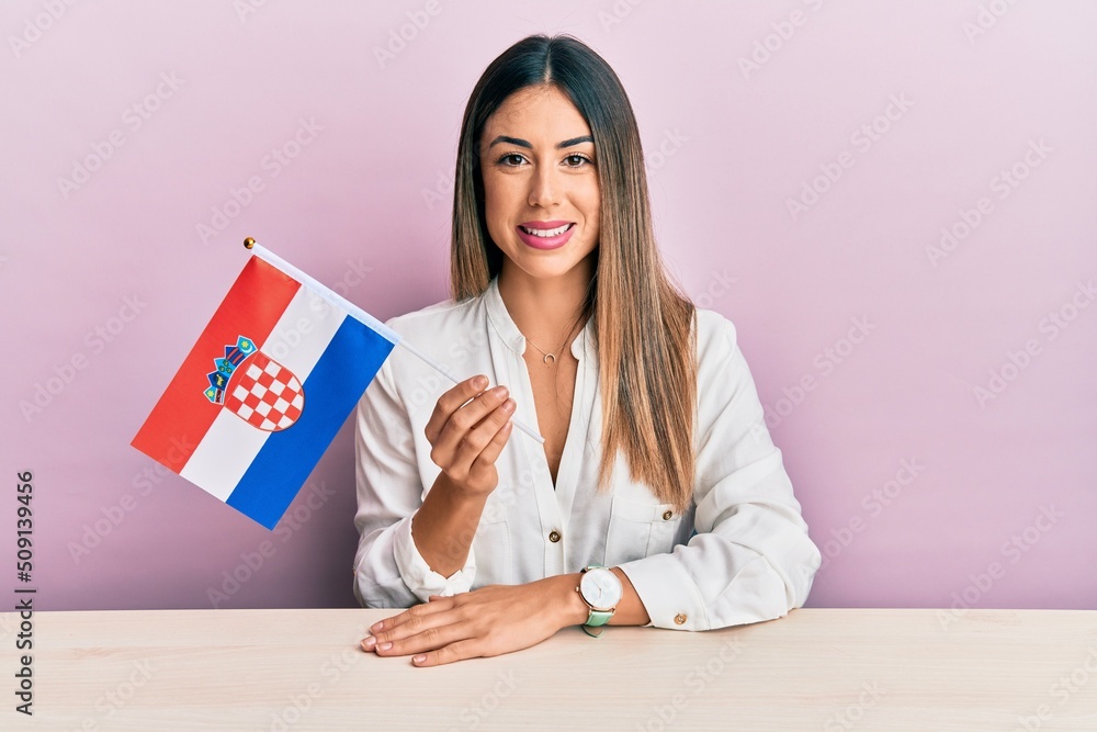 Young hispanic woman holding croatia flag sitting on the table looking positive and happy standing and smiling with a confident smile showing teeth Young hispanic woman holding croatia flag sitting on the table looking positive and happy standing and smiling with a confident smile showing teeth