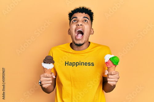 Young african american man wearing t shirt with happiness word message holding ice cream angry and mad screaming frustrated and furious, shouting with anger looking up.