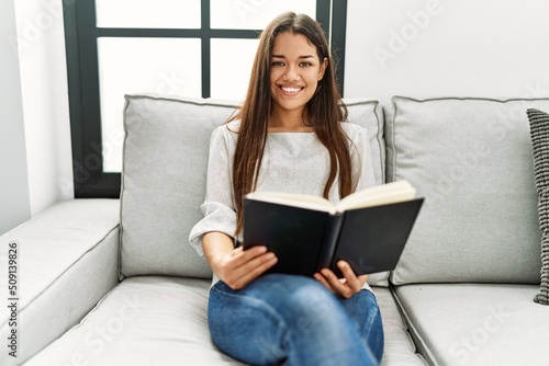 Young latin woman reading book sitting on sofa at home