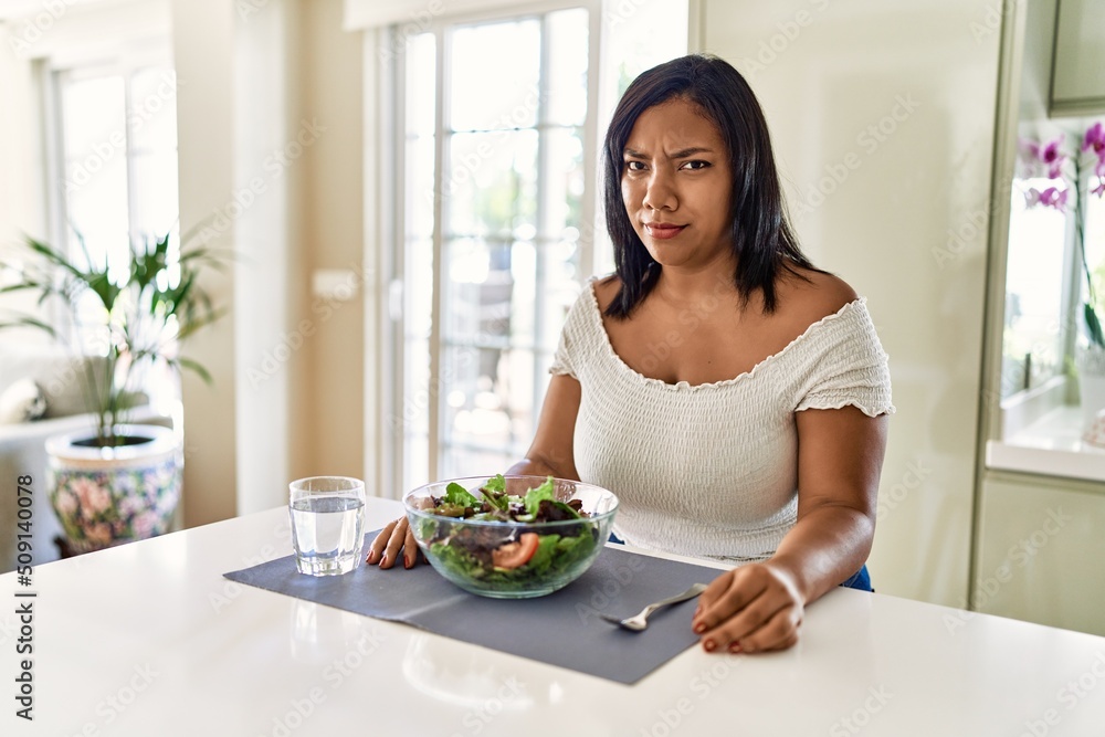 Young hispanic woman eating healthy salad at home skeptic and nervous, frowning upset because of problem. negative person.