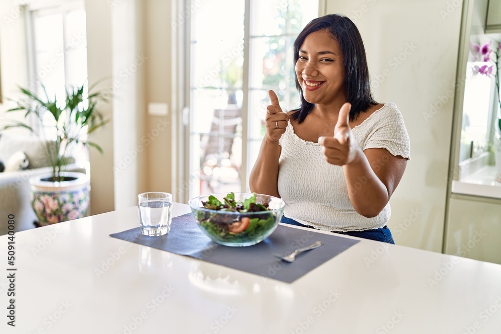Young hispanic woman eating healthy salad at home pointing fingers to camera with happy and funny face. good energy and vibes.