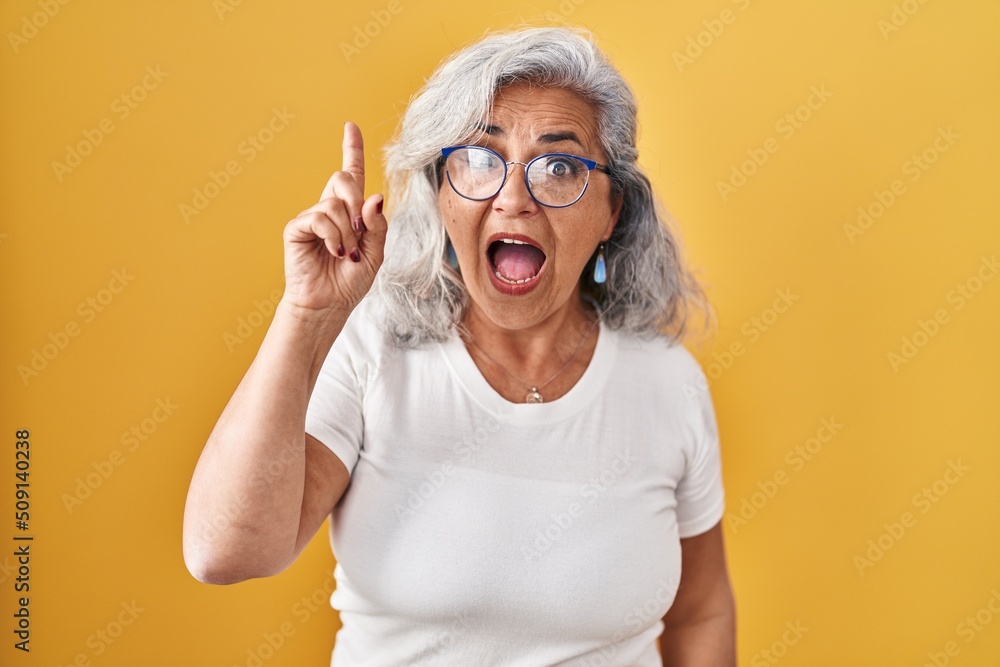 Middle age woman with grey hair standing over yellow background pointing finger up with successful idea. exited and happy. number one. Middle age woman with grey hair standing over yellow background pointing finger up with successful idea. exited and happy. number one.