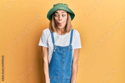 Young caucasian blonde woman wearing denim jumpsuit and hat with 90s style making fish face with lips, crazy and comical gesture. funny expression.