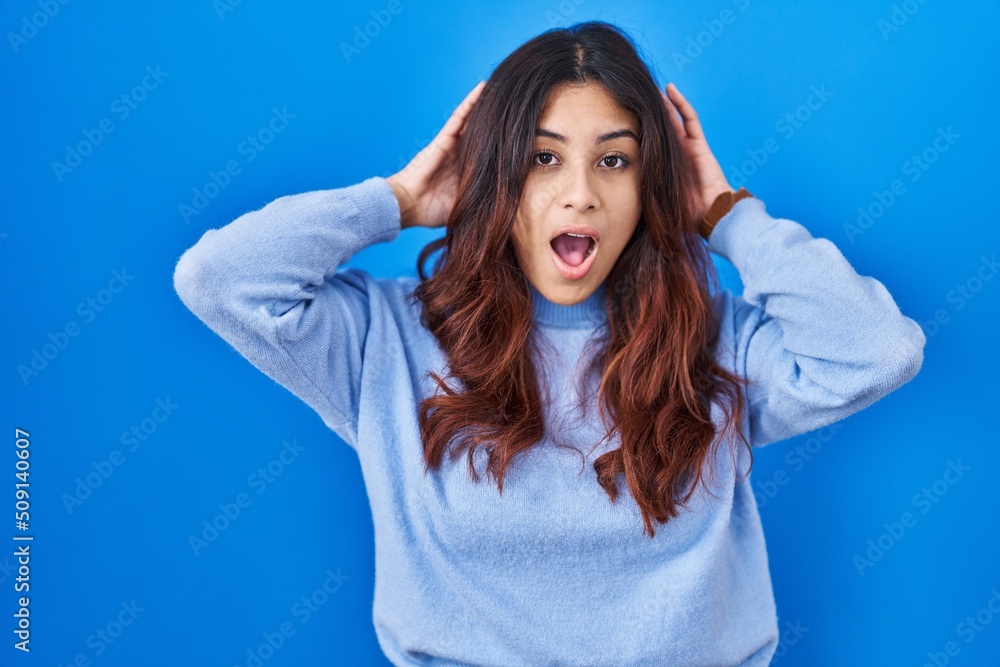 Hispanic young woman standing over blue background crazy and scared with hands on head, afraid and surprised of shock with open mouth
