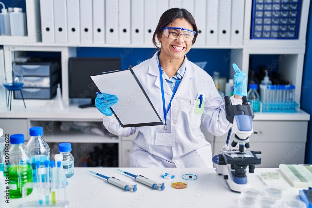 Hispanic young woman working at scientist laboratory very happy and excited doing winner gesture with arms raised, smiling and screaming for success. celebration concept.