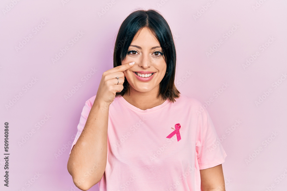 Young hispanic woman wearing pink cancer ribbon on t shirt pointing with hand finger to face and nose, smiling cheerful. beauty concept Young hispanic woman wearing pink cancer ribbon on t shirt pointing with hand finger to face and nose, smiling cheerful. beauty concept