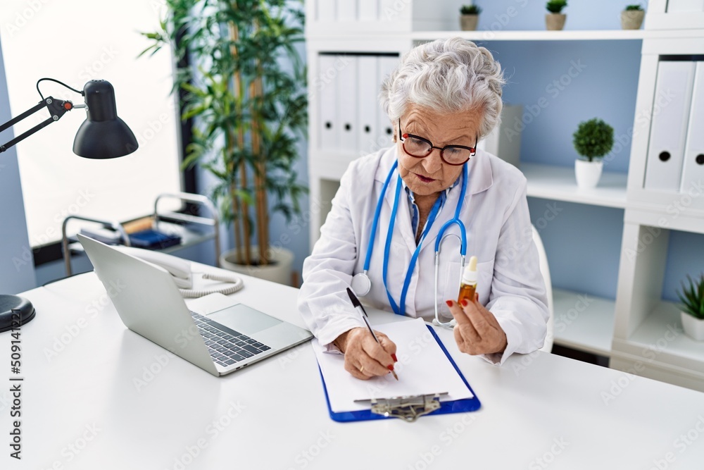Senior grey-haired woman wearing doctor uniform writing on clipboard holding treatment at clinic