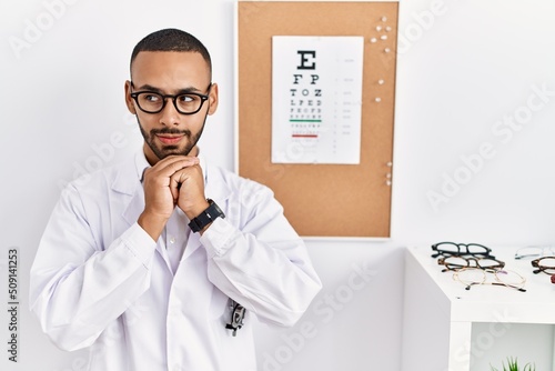 African american optician man standing by eyesight test laughing nervous and excited with hands on chin looking to the side