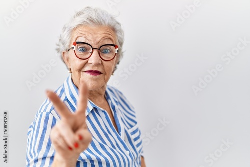 Senior woman with grey hair standing over white background smiling looking to the camera showing fingers doing victory sign. number two.
