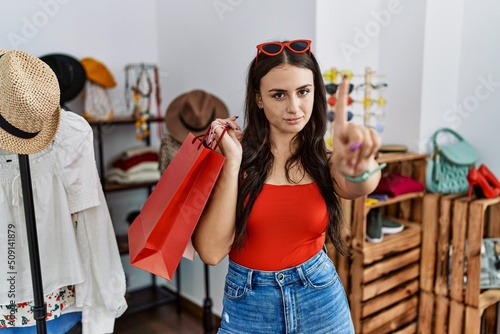 Young brunette woman holding shopping bags at retail shop pointing with finger up and angry expression, showing no gesture