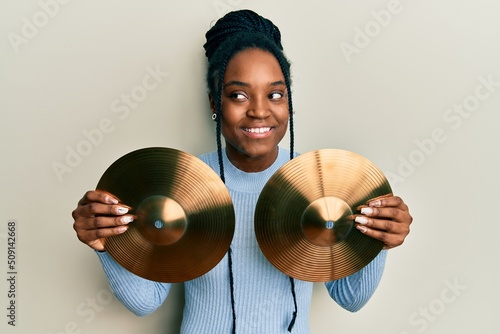 African american woman with braided hair holding golden cymbal plates smiling looking to the side and staring away thinking.