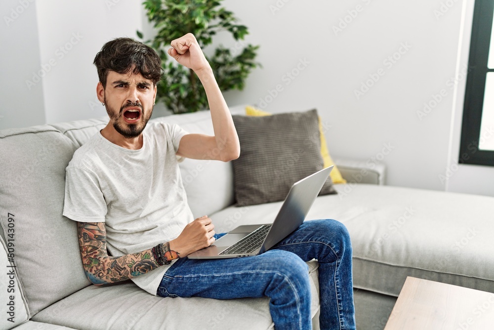 Hispanic man with beard sitting on the sofa angry and mad raising fist frustrated and furious while shouting with anger. rage and aggressive concept. Hispanic man with beard sitting on the sofa angry and mad raising fist frustrated and furious while shouting with anger. rage and aggressive concept.