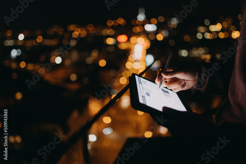 Close-up of man standing on balcony with urban view and using tablet at night