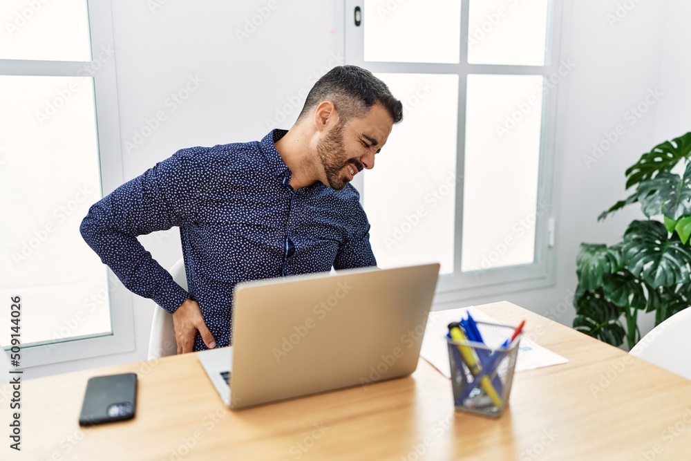 Young hispanic man with beard working at the office with laptop suffering of backache, touching back with hand, muscular pain Young hispanic man with beard working at the office with laptop suffering of backache, touching back with hand, muscular pain