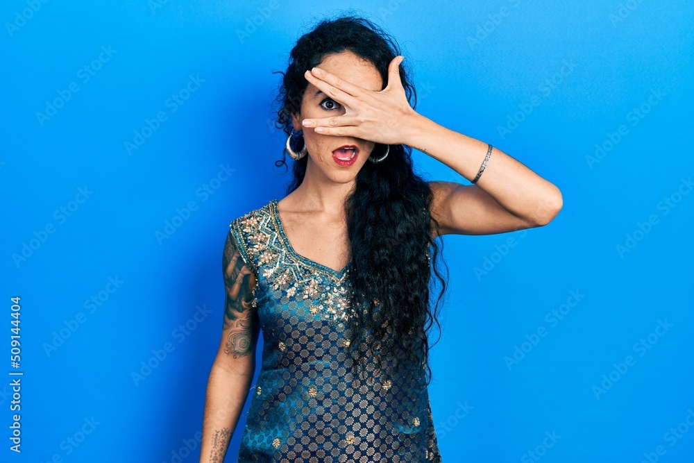 Young woman wearing bindi and traditional kurta dress peeking in shock covering face and eyes with hand, looking through fingers with embarrassed expression.