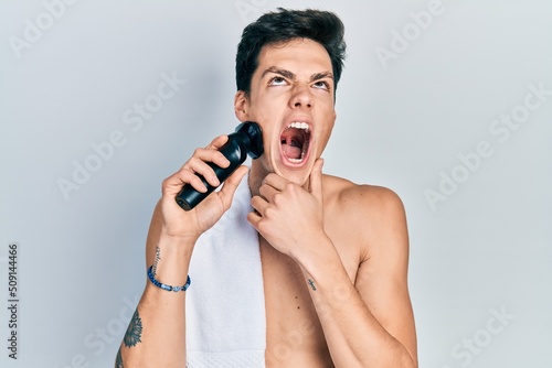 Young hispanic man shaving with electric razor machine angry and mad screaming frustrated and furious, shouting with anger looking up.