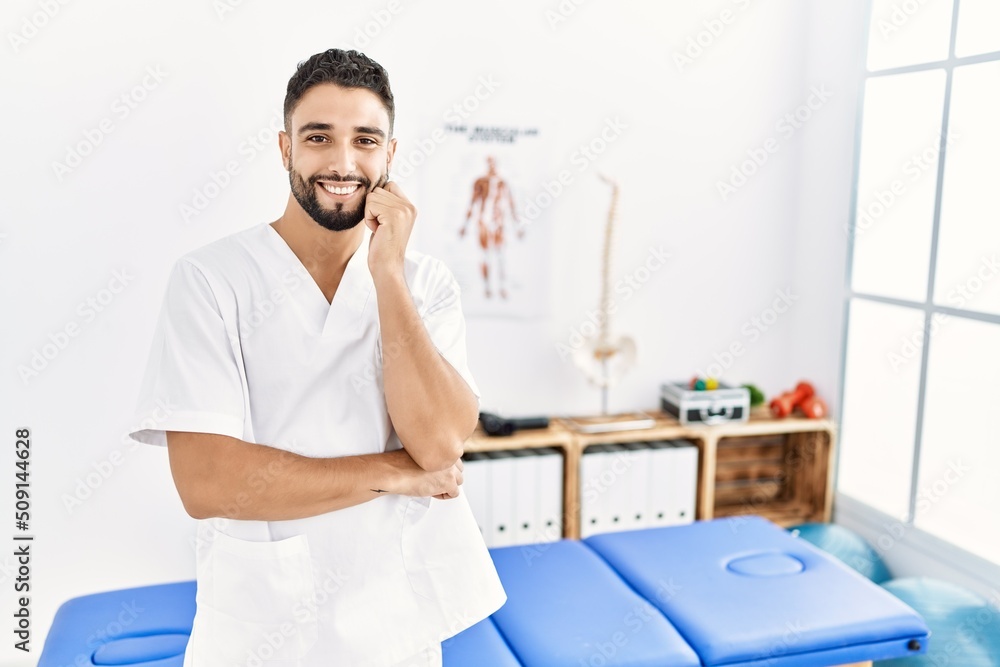 Young handsome man with beard working at pain recovery clinic looking confident at the camera smiling with crossed arms and hand raised on chin. thinking positive. Young handsome man with beard working at pain recovery clinic looking confident at the camera smiling with crossed arms and hand raised on chin. thinking positive.