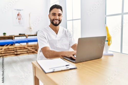 Young arab man wearing physiotherapist uniform using laptop at clinic