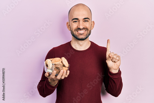 Young bald man holding sweet pastries smiling with an idea or question pointing finger with happy face, number one