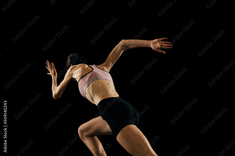 Back view of young muscular woman running isolated on black background. Sport, track-and-field athletics, competition and active lifestyle concept