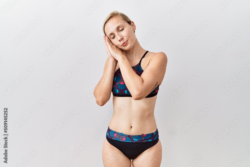 Young blonde woman wearing sporty bikini over isolated background sleeping tired dreaming and posing with hands together while smiling with closed eyes. Young blonde woman wearing sporty bikini over isolated background sleeping tired dreaming and posing with hands together while smiling with closed eyes.