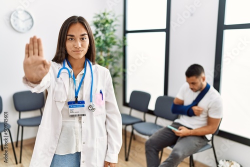 Young asian doctor woman at waiting room with a man with a broken arm doing stop sing with palm of the hand. warning expression with negative and serious gesture on the face.