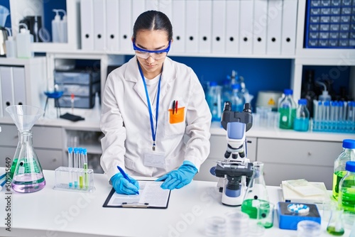 Young hispanic woman scientist writing on document working at laboratory
