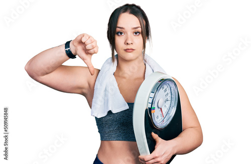 Young hispanic girl wearing sportswear holding weighing machine with angry face  negative sign showing dislike with thumbs down  rejection concept