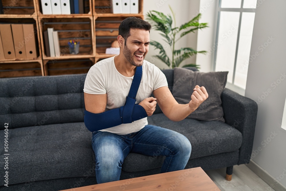 Young hispanic man with beard wearing arm on sling sitting at therapy consult annoyed and frustrated shouting with anger, yelling crazy with anger and hand raised Young hispanic man with beard wearing arm on sling sitting at therapy consult annoyed and frustrated shouting with anger, yelling crazy with anger and hand raised