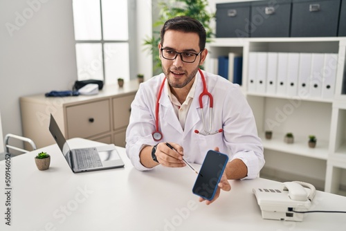 Wallpaper Mural Young hispanic man wearing doctor uniform showing medical smartphone app at clinic Torontodigital.ca