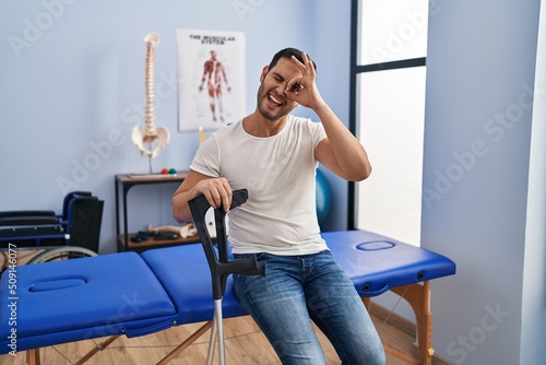 Young hispanic man with beard wearing crutches at rehabilitation clinic smiling happy doing ok sign with hand on eye looking through fingers