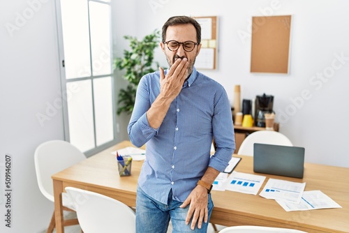 Middle age hispanic man with beard wearing business clothes at the office bored yawning tired covering mouth with hand. restless and sleepiness. photo